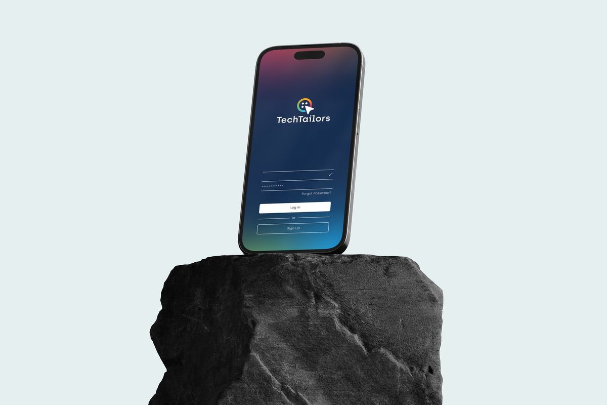 A phone standing on a dark rock, its sign-in screen showing the TechTailors button mark and name.